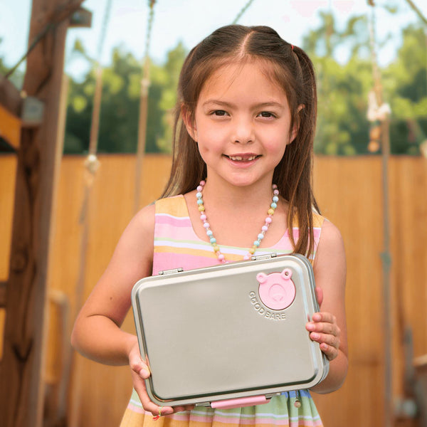 Young girl holding a stainless steel lunchbox with a pink bear emblem outdoors.