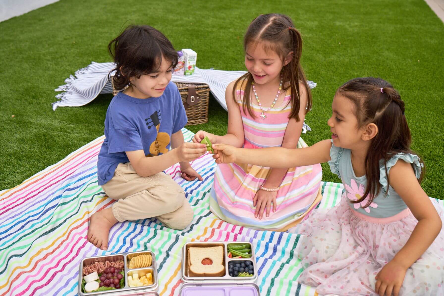 Three children sitting on a colorful blanket with picnic food, enjoying a sunny day outdoors with Good Bare stainless steel lunch boxes