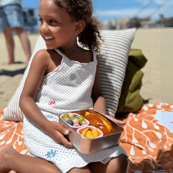 A girl sitting on a beach blanket holding a stainless steel sandwich box with food.