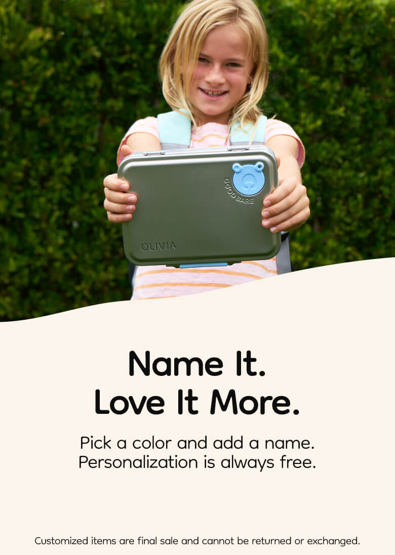 Child holding a stainless steel lunch box with a brand logo against a green hedge background, featuring the text 'Name It. Love It More.